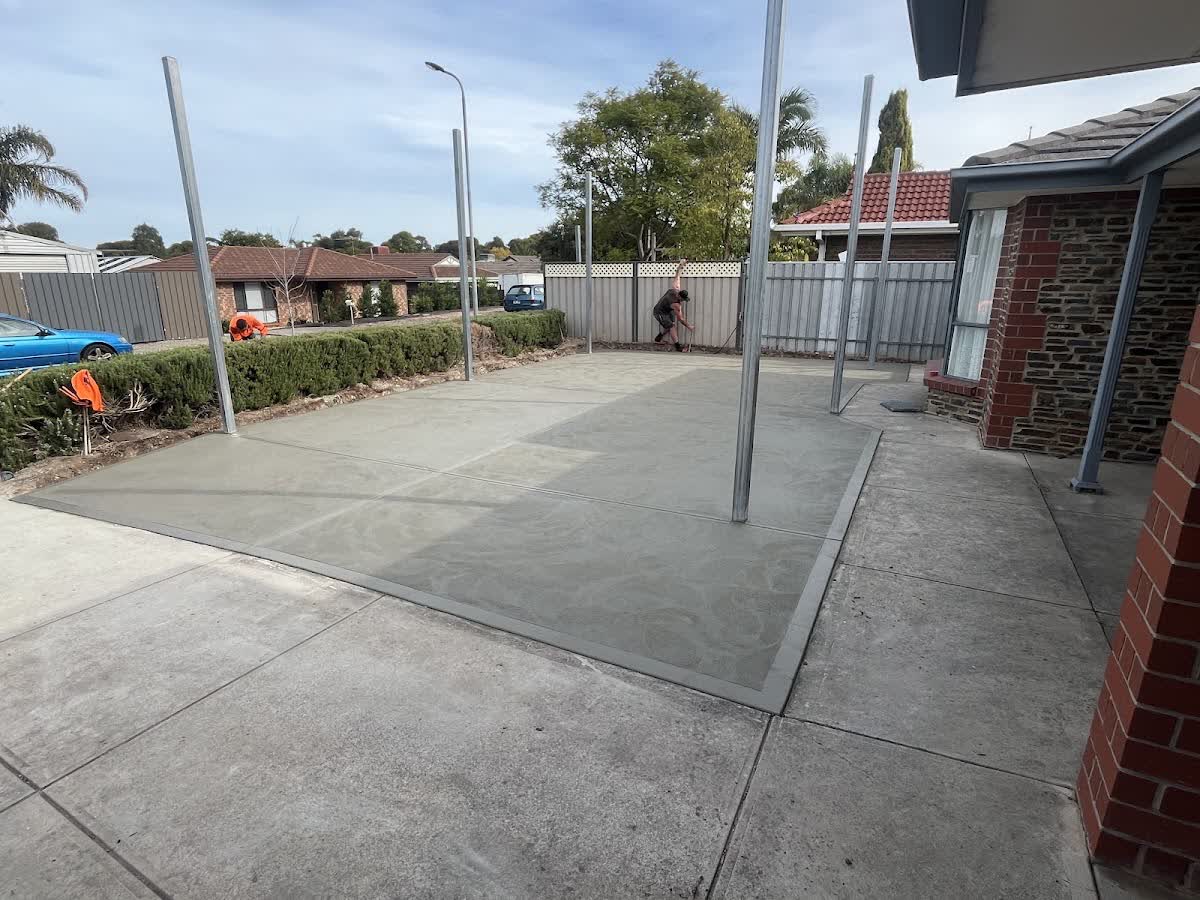 A large scored broom-finish concrete patio slab poured under a carport, the crew finishing the edge