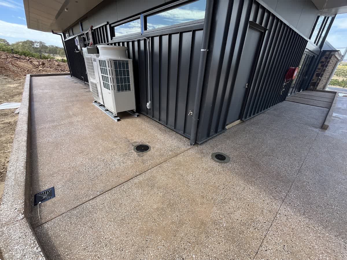 A broad exposed-aggregate concrete slab with drainage points beside a modern build